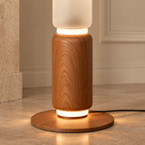 A modern table lamp with a cylindrical wooden body, a frosted white top section, and a round wooden base that glows with warm light, set on a stone floor in an indoor setting.