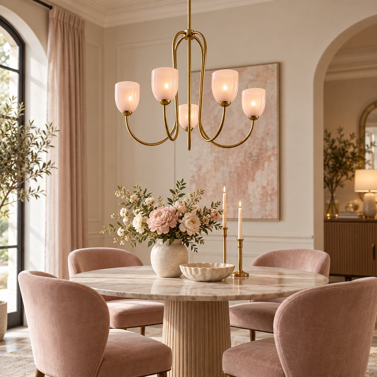 A modern brass chandelier with six glowing warm-white glass shades hangs over a round marble dining table in a styled dining room with blush pink velvet chairs, a floral centerpiece, and soft neutral walls.
