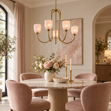 A modern brass chandelier with six glowing warm-white glass shades hangs over a round marble dining table in a styled dining room with blush pink velvet chairs, a floral centerpiece, and soft neutral walls.