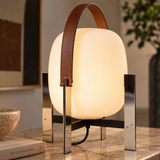 A modern table lamp with a glowing oval white shade, a brown leather carry handle, and a polished silver metal stand, placed on a marble tabletop in a styled interior setting.