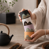 A person holds a stack of four ceramic tea cups with mountain-patterned rims in soft pastel colors. The setting is a warm, styled tea table scene with a black teapot, loose leaf dish, and neutral fabric background.