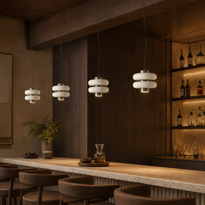 Three modern pendant lights with stacked white cylindrical shades hang over a wooden bar counter in a warm, dark-toned interior with bar stools and shelving.