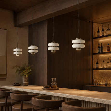 Three modern pendant lights with stacked white cylindrical shades hang over a wooden bar counter in a warm, dark-toned interior with bar stools and shelving.