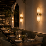 A warm wall-mounted sconce with a vertical frosted glass shade and gold-toned metal trim, installed on a restaurant wall above a booth and dining tables.