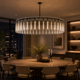 A large circular chandelier with clear textured glass and warm vertical light tubes, hanging over a round wooden dining table in a modern, warmly lit dining room.