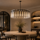 A large modern chandelier with a circular frame and hanging clear glass or crystal-like drops, glowing with warm light above a round dining table in a styled interior room.