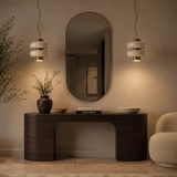 A modern dark wood console table in a warm, minimalist interior setting, styled with a dark ceramic vase, a small bowl, a book, and a white ceramic bowl. Above the table is a large, vertically-oriented oval mirror, flanked by two hanging pendant lights with white stacked glass shades and gold accents.