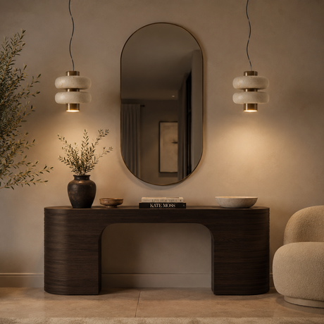 A modern dark wood console table in a warm, minimalist interior setting, styled with a dark ceramic vase, a small bowl, a book, and a white ceramic bowl. Above the table is a large, vertically-oriented oval mirror, flanked by two hanging pendant lights with white stacked glass shades and gold accents.