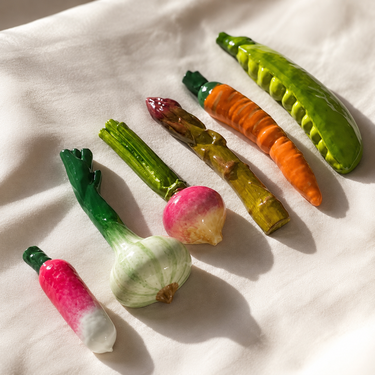 Harvest Artisan vegetable-shaped ceramic cutlery rests arranged on cream fabric in warm natural light.
