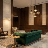 A luxury modern lobby featuring a plush emerald green velvet tufted sofa, a round marble-top side table with a gold brass lamp, a floor lamp, and a multi-light glass chandelier against dark wood paneling and beige stone walls.