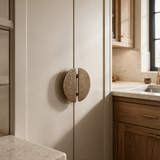 A pair of round, textured bronze cabinet handles mounted on cream-colored kitchen cabinets, with a modern kitchen sink and wood cabinetry visible in the background.