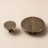 Two round cabinet knobs in an antique brass finish with a textured, wood-grain-like surface, displayed on a warm beige background.