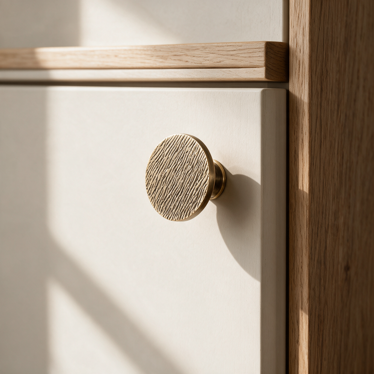 A close-up of a round, textured brass cabinet knob mounted on a white drawer front, set within a light wood frame under warm natural light.