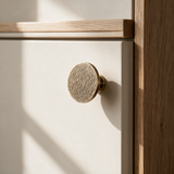 A close-up of a round, textured brass cabinet knob mounted on a white drawer front, set within a light wood frame under warm natural light.