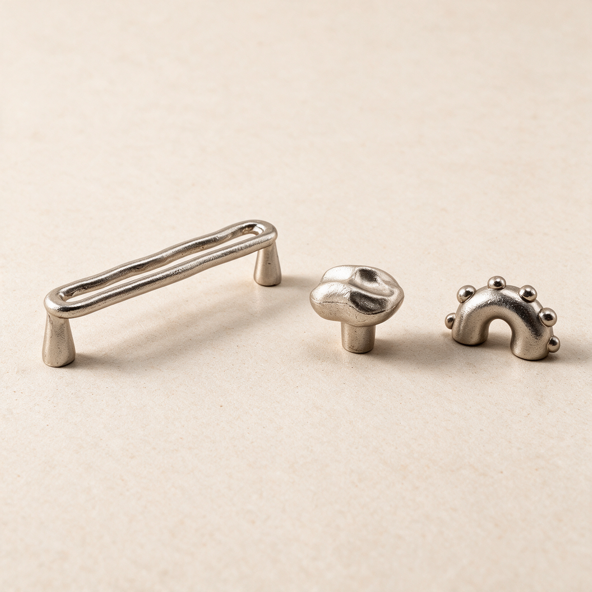 Three small metallic cabinet hardware pieces on a warm beige background: a long rectangular pull handle, a round knob, and a small decorative bracket with raised dots.