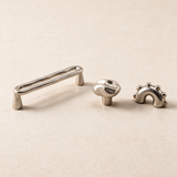 Three small metallic cabinet hardware pieces on a warm beige background: a long rectangular pull handle, a round knob, and a small decorative bracket with raised dots.