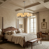 A luxury bedroom featuring a dark wood four-poster bed with beige linens, a matching wooden bench at the foot of the bed, a modern multi-light chandelier, and a cozy desk setup by a window.