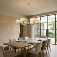 A modern multi-light chandelier with frosted oval glass shades hangs over a wooden dining table and upholstered chairs in a bright, contemporary dining room with stone walls and large windows.