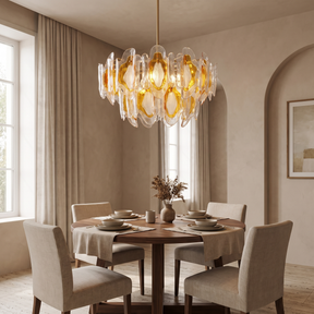 A modern multi-light chandelier with amber and clear glass leaf-like elements hangs over a round wooden dining table with four upholstered chairs in a warm, neutral-toned dining room.
