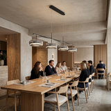 A long wooden dining table with light wood chairs in a modern, warm-toned conference or dining room. Several people are seated around the table, and the space is illuminated by a linear pendant light with multiple silver and black shades.