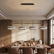 A modern 4-light linear ceiling pendant chandelier with textured gray and silver drum shades, suspended over a wooden dining table in a contemporary dining room.