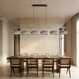 A modern 6-light linear pendant chandelier with brushed metal shades hangs over a dining table and chairs in a minimalist, warm-toned dining room with a textured beige stone wall.