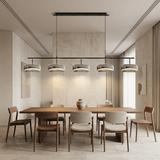 A modern 5-light linear pendant chandelier with brushed silver metal shades and black suspension hardware, hanging above a wooden dining table and chairs in a minimalist, warm-toned dining room.