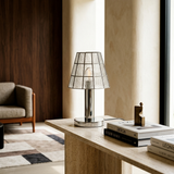 A modern table lamp with a textured clear glass conical shade and a polished silver metal base, styled on a light stone console table in a warm, contemporary living room.