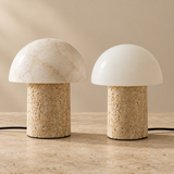 Two mushroom-shaped table lamps on a stone surface against a warm beige background. One lamp has a cream marble dome shade and the other has a smooth white dome shade, both with textured cylindrical bases.