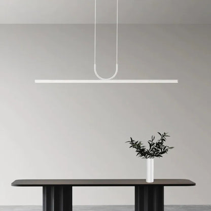 A minimalist white linear pendant light hanging above a modern black dining table, with a simple vase of greenery placed on the table. The soft light illuminates a neutral-colored wall in the background.