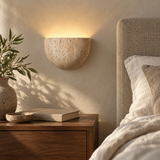Lumo Travertine Wall Lamp casting warm uplight above a walnut bedside table beside a linen headboard.
