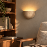 Lumo Travertine Wall Lamp with warm uplight beside walnut shelves and a linen armchair in a reading nook.