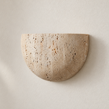 Close-up of the Lumo Travertine Wall Lamp switched off, showing its rounded shape and porous beige stone on a plaster wall.