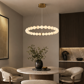 A modern ring pendant light with spherical glass shades hangs over a round dining table in a styled interior with chairs, dark wood cabinetry, and a vase of greenery.