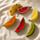 Five colorful fruit slices arranged on a draped cream fabric background: pineapple, red watermelon, yellow lemon, orange melon, and kiwi.