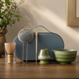 A tea preparation set arranged on a wooden table in a softly lit interior. The set includes a blue zippered case with a handle, a metal mesh strainer, a bamboo whisk in a clear glass holder, a small green pitcher, and a green bowl.