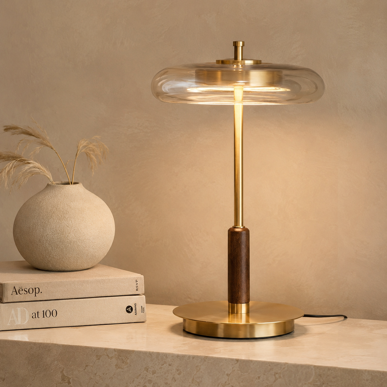 A modern table lamp with a round clear glass shade, a brushed brass stem and base, and a dark wood accent, shown lit on a stone surface next to stacked books and a vase.