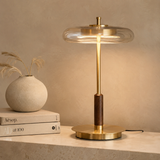 A modern table lamp with a round clear glass shade, a brushed brass stem and base, and a dark wood accent, shown lit on a stone surface next to stacked books and a vase.