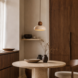 A modern pendant light with a warm glowing dome shade and a round wooden accent, hanging over a round stone dining table in a minimalist, warm-toned interior.
