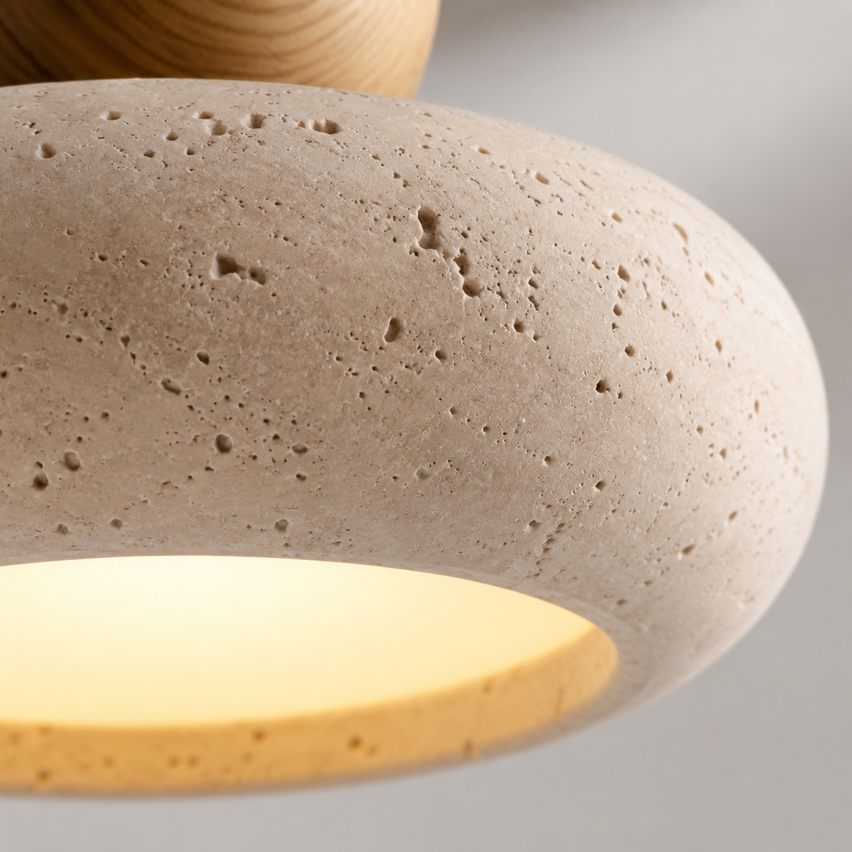 Close-up of a modern pendant light with a textured beige travertine stone shade and a warm yellow illuminated interior, with a wooden finial visible at the top.