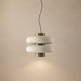 A modern two-tier pendant light fixture with textured off-white stone rings and brushed brass metal accents, suspended against a warm beige textured wall.