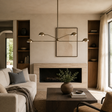 A modern living room interior with a beige sofa, dark wood coffee table, built-in wood shelving, a fireplace, and a brass multi-arm pendant light hanging above a framed abstract print.