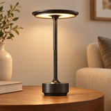 A modern table lamp with a circular black metal shade and a textured black stem stands on a round wooden table in a warm, cozy living room setting.