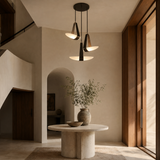 Three modern pendant lights with black metal frames and warm glowing shades hang over a round stone table in a minimalist, warm-toned interior with beige walls and wood accents.