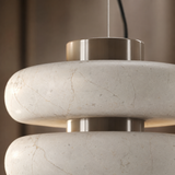 Close-up of a modern pendant light with thick, rounded off-white stone or marble discs and brushed metallic silver-toned rings, suspended by a thin cord against a warm brown background.