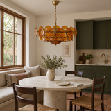 Unlit Velari Disc Orange Glass Chandelier above a round stone breakfast table beside moss-green kitchen cabinets and a timber-framed window.
